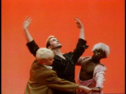 Three dancers perform in front of a red background. One dancer stretches their arms to the sky, the other dancers contain the first within their arms.